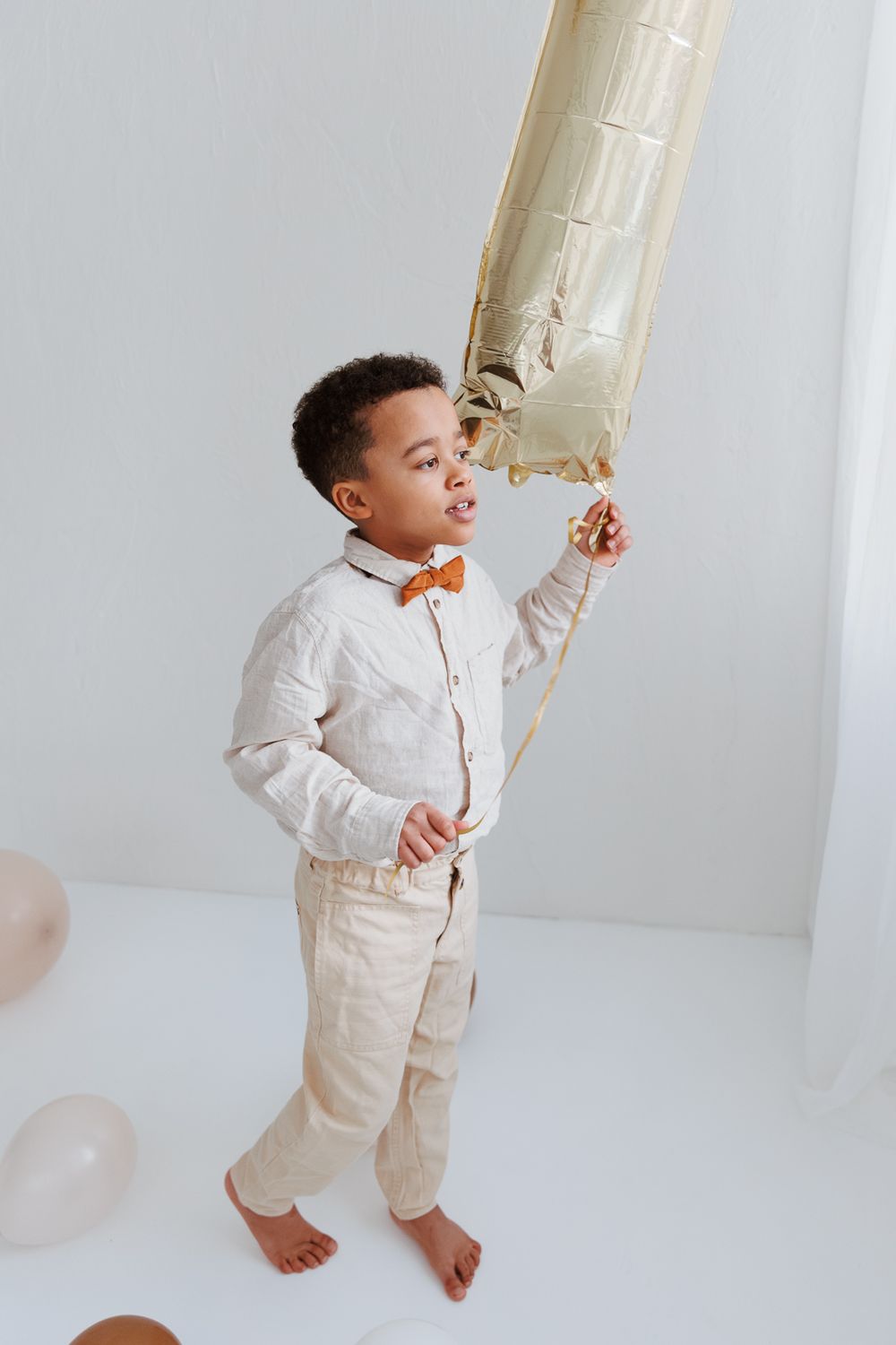 Young child in formal cream outfit with orange bow tie holds a large decorative balloon in a bright minimalist setting.