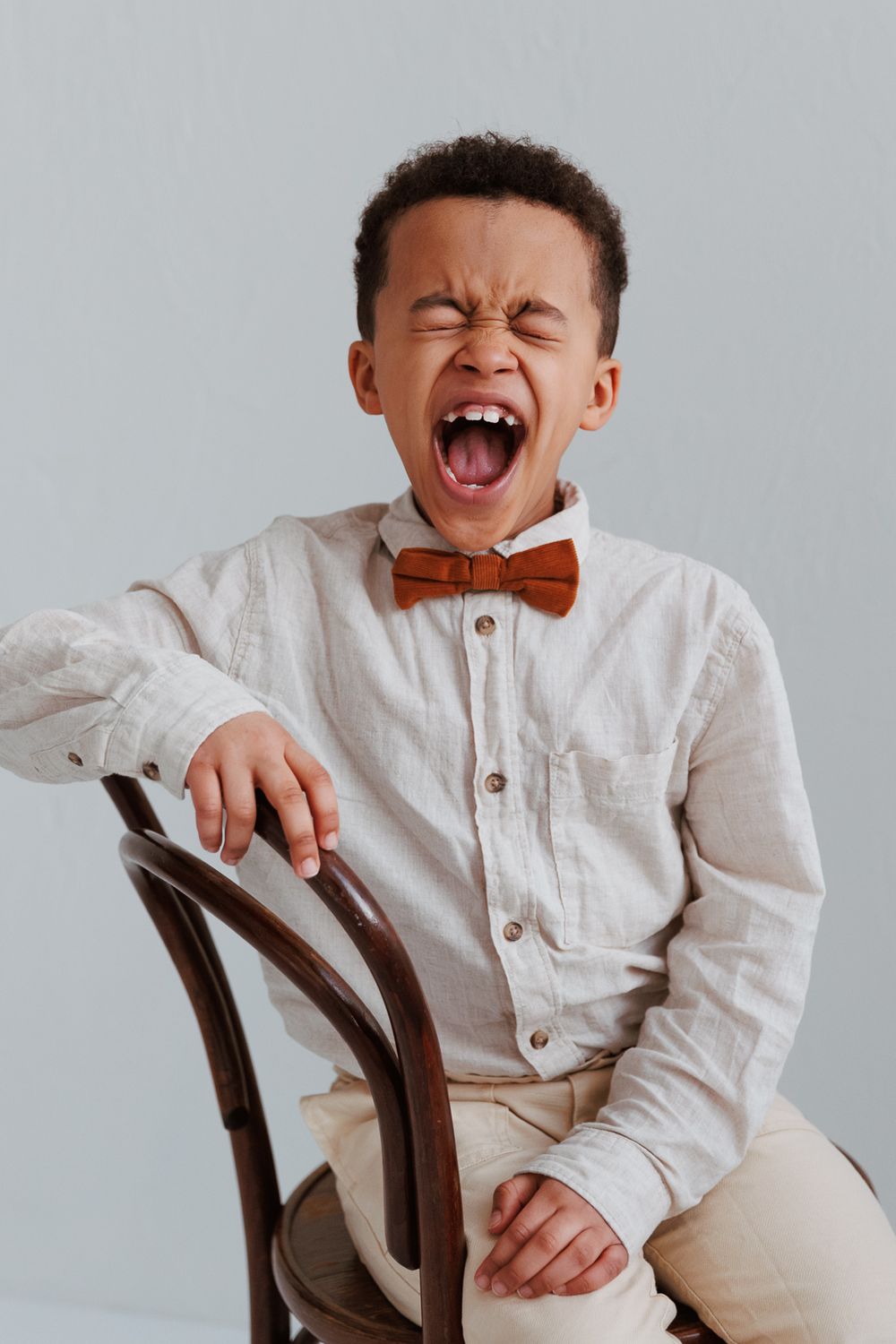 Young child in white shirt and brown bowtie sitting on chair with exuberant yawning expression against light background.