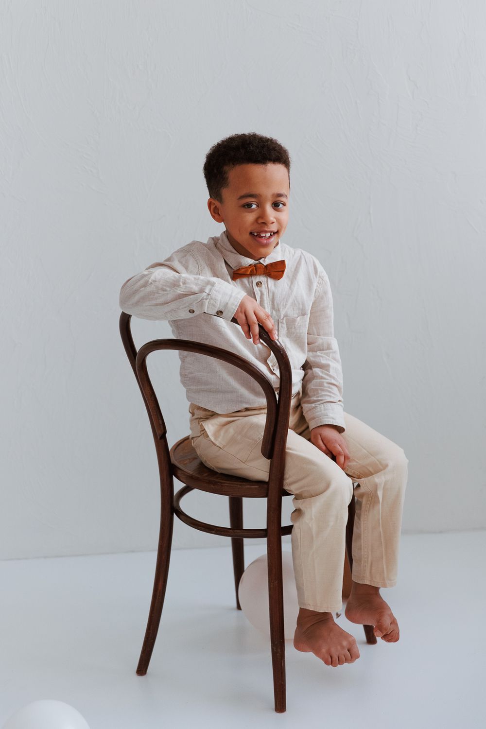 Child in white outfit poses playfully on wooden chair against light background in minimalist photo series.