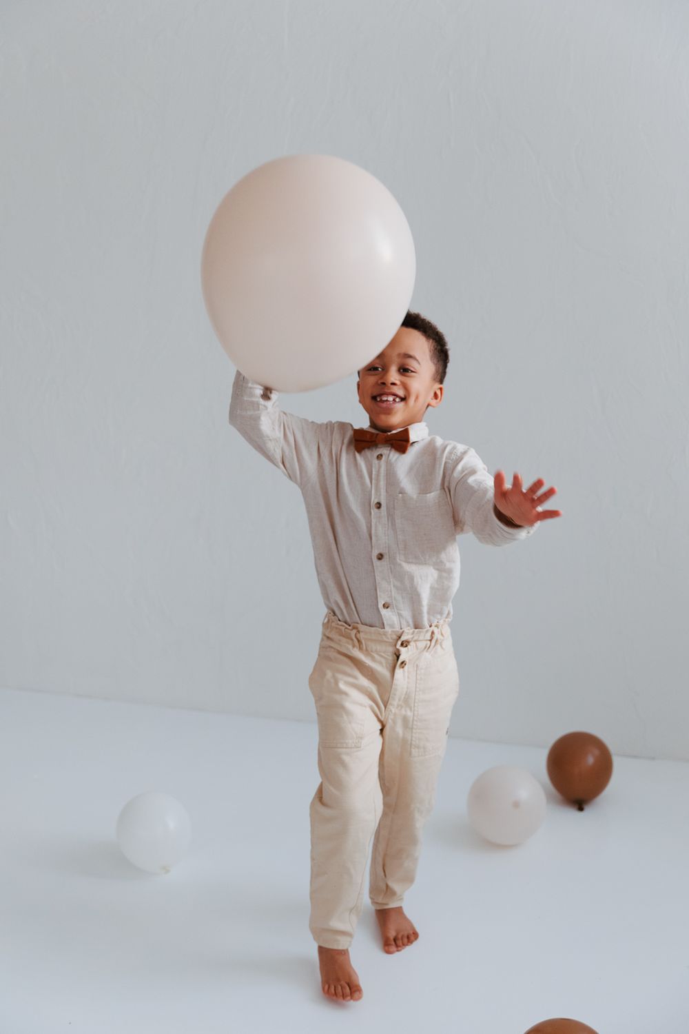 A person in white and beige outfit playfully interacts with a balloon in a minimalist white studio setting.
