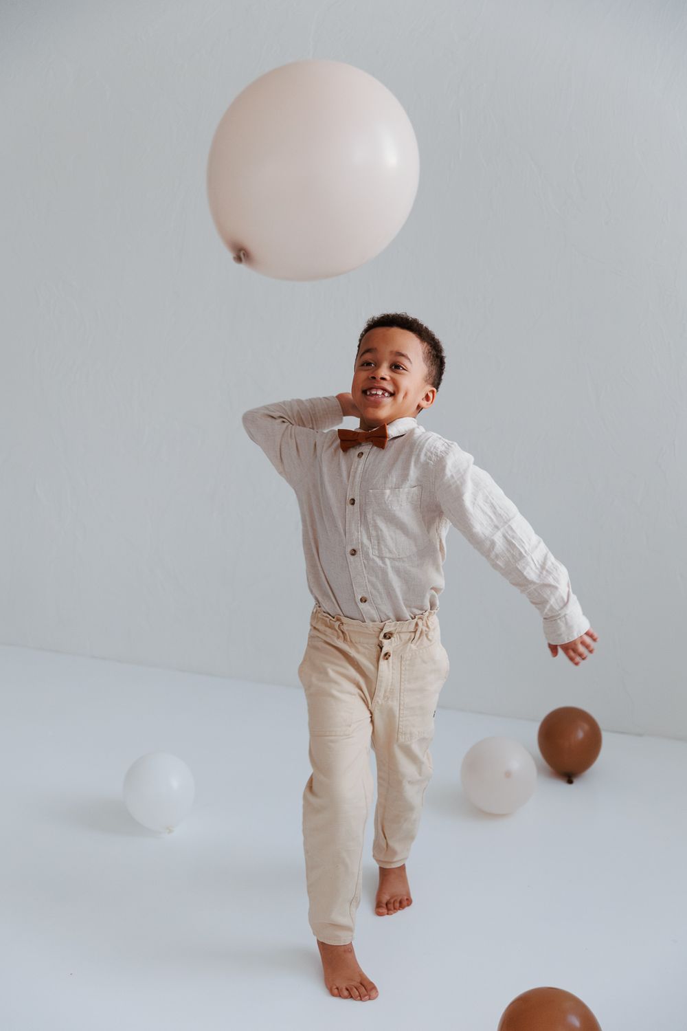 Young child in formal attire playing with balloons on white background while barefoot.