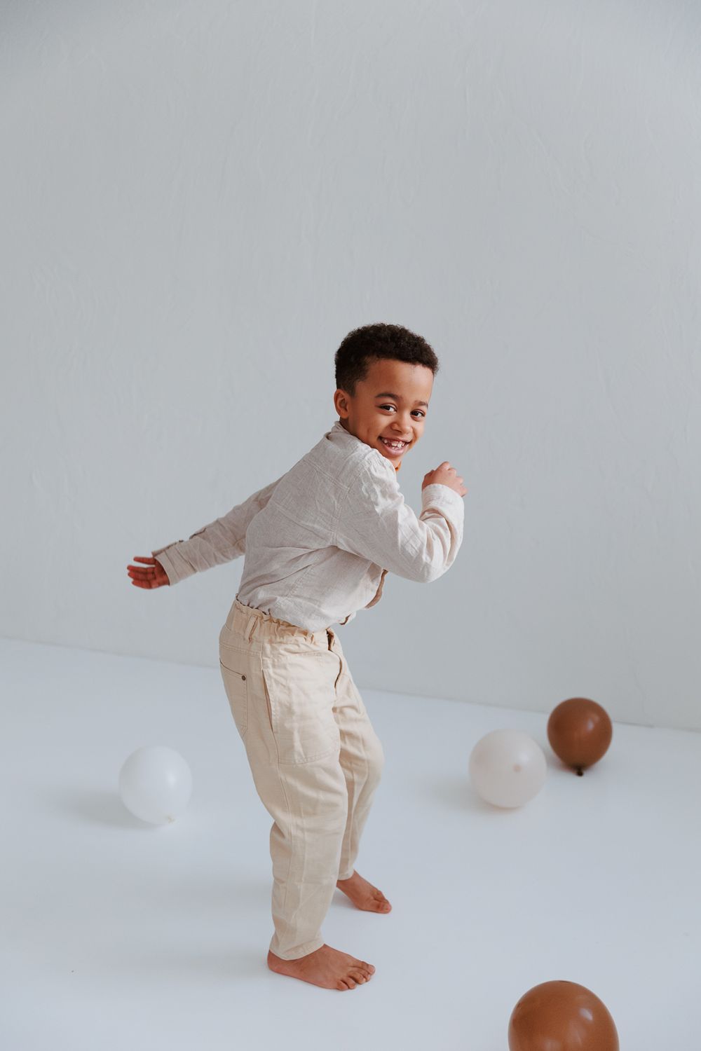 Child in white shirt and beige pants dancing playfully with balloons on white floor.