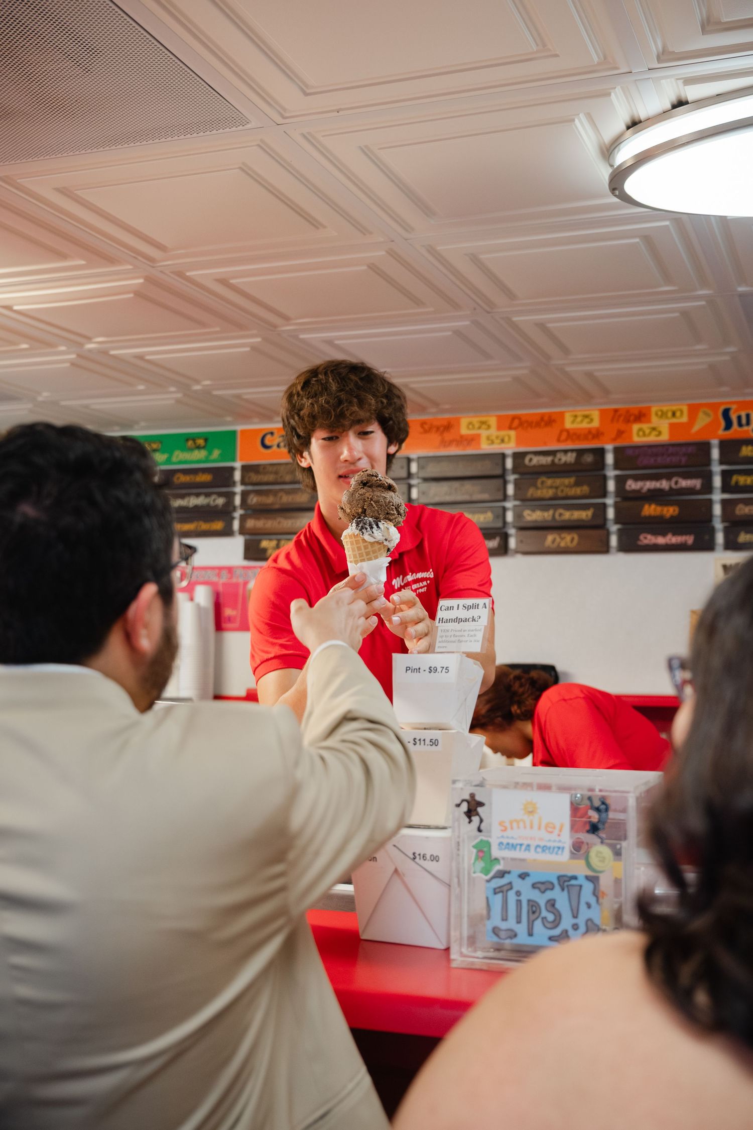 An ice cream shop employee in a red uniform serves a customer at the counter while others wait in line.