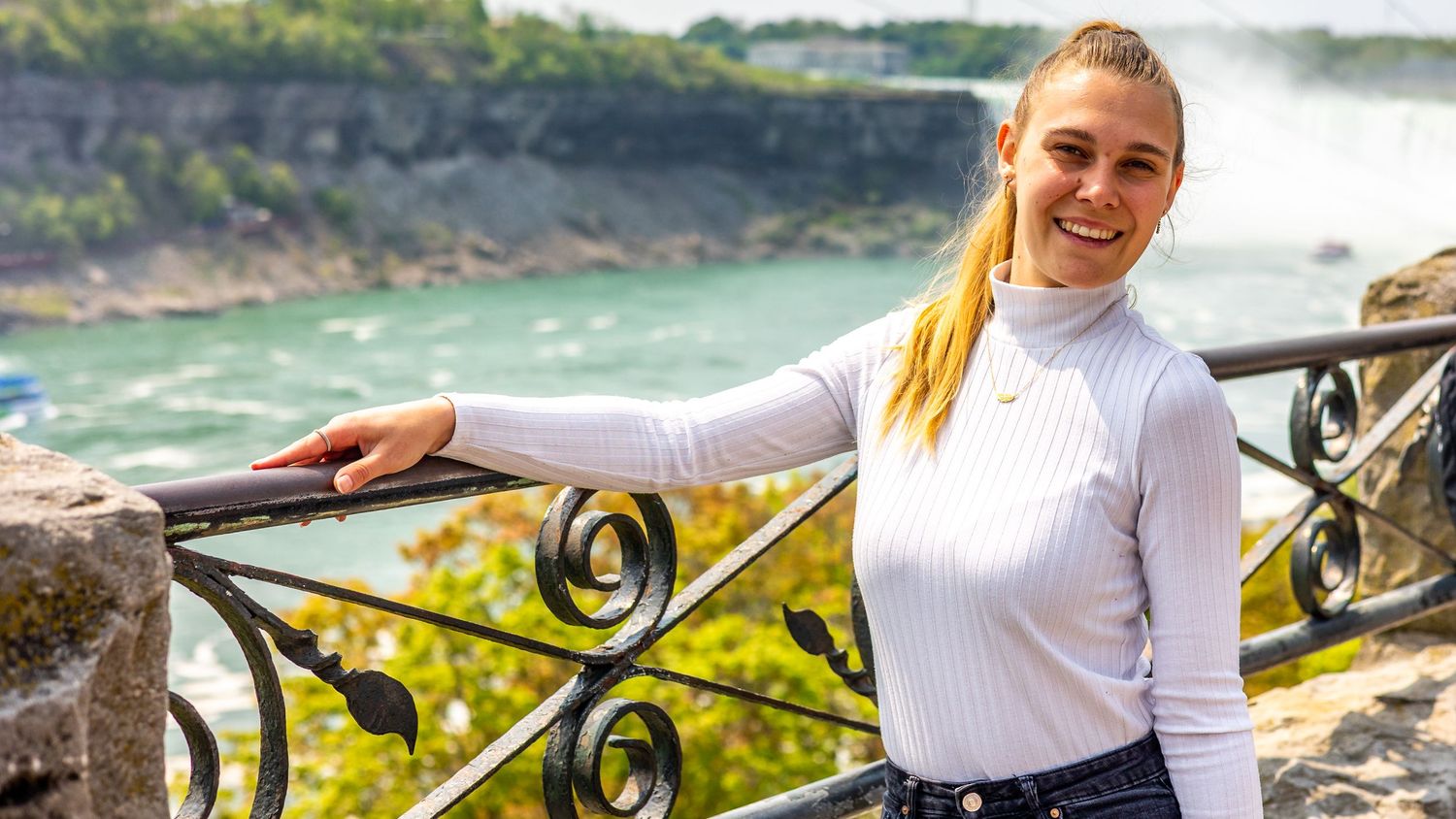A person in a white sweater poses near a scenic waterfall overlook with ornate metal railings in the background.