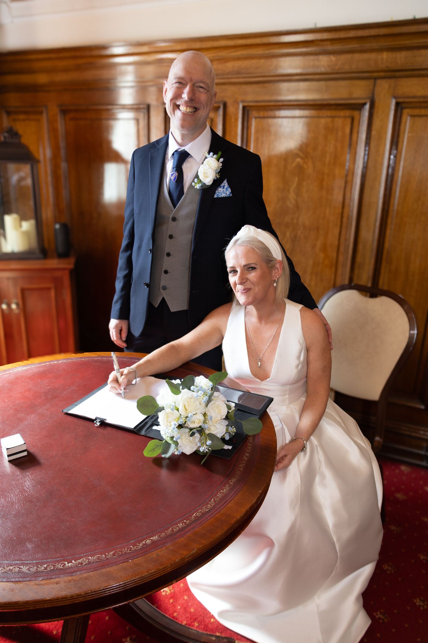 A couple signs documents at an elegant wooden table during their wedding ceremony with white floral bouquet nearby.