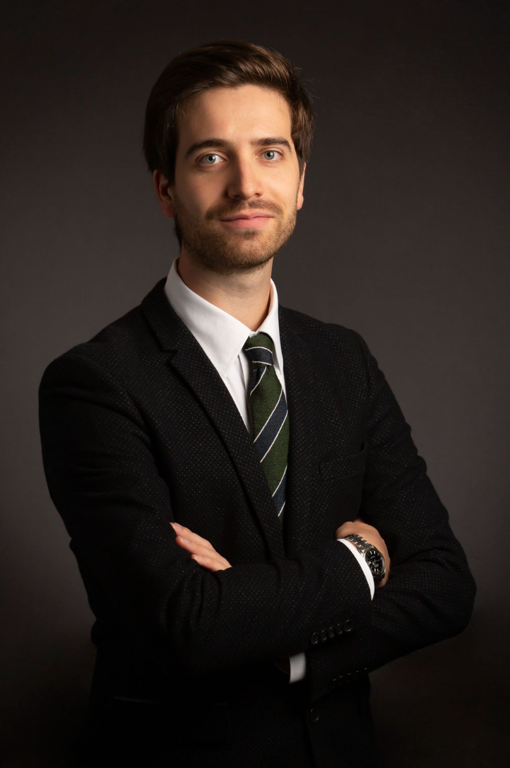 Professional headshot of a person in a dark suit with striped tie against a dark background, arms crossed confidently.