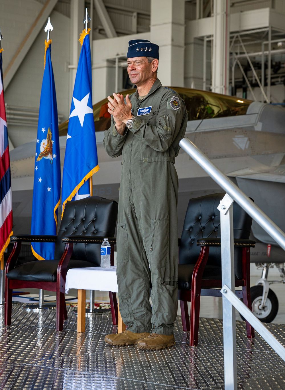 Military officer in flight suit applauds at ceremony in aircraft hangar with US Air Force flags and jet in background.
