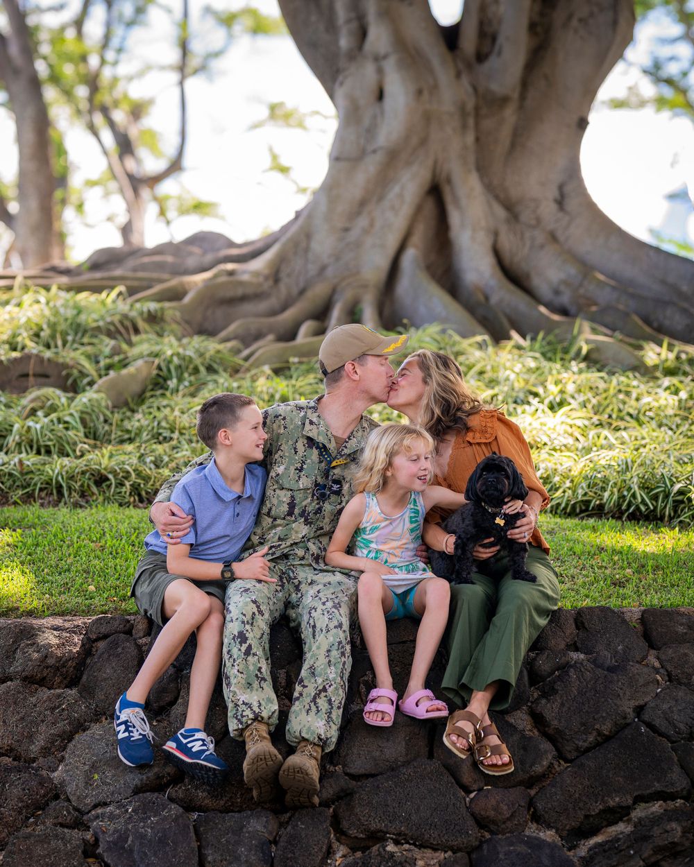 Military family with two children and a puppy sharing a tender moment on a stone wall beneath a large banyan tree.