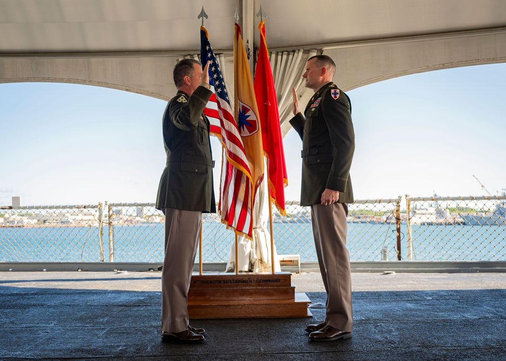 Two military officers in dress uniforms stand at attention with American and Army flags during a formal ceremony outdoors.