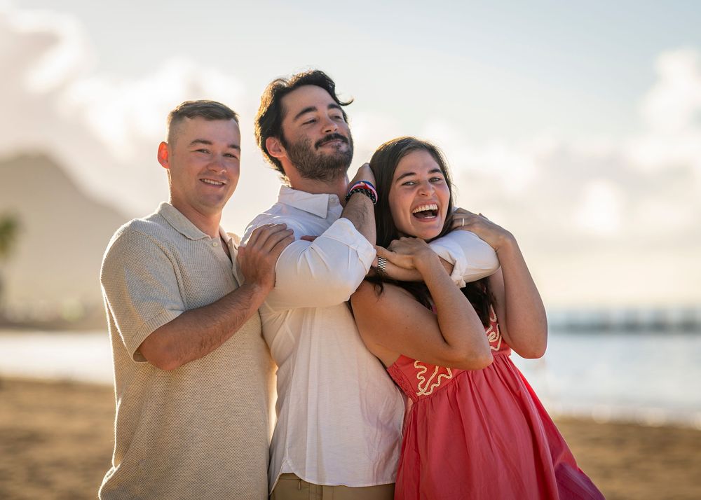 Three friends laughing and embracing on a tropical beach at sunset, wearing casual summer attire.