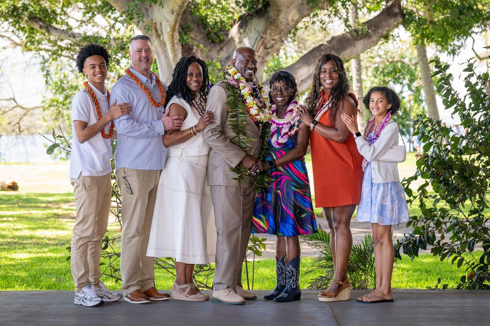 A joyful group of six people posing outdoors under a large tree, wearing festive leis and colorful attire.
