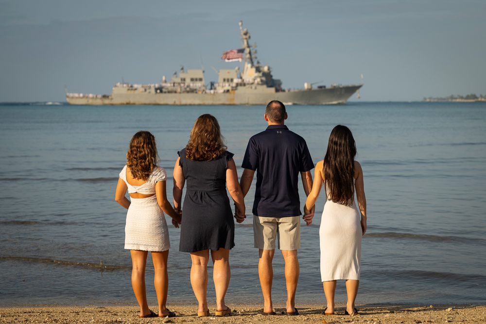 Family portraits Oahu at Pearl Harbor entrance at beach's edge watching the ocean and U.S. Navy ship at sunrise.