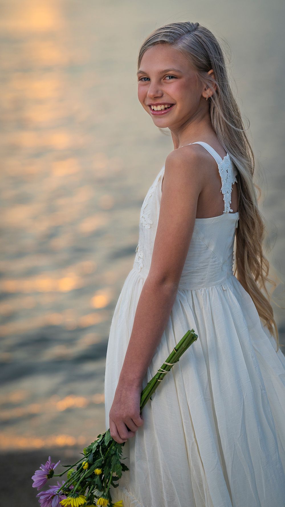 Your girl in a white dress holds flowers by the Pacific Ocean and water's edge at sunset, creating a serene childhood portrait in Honolulu