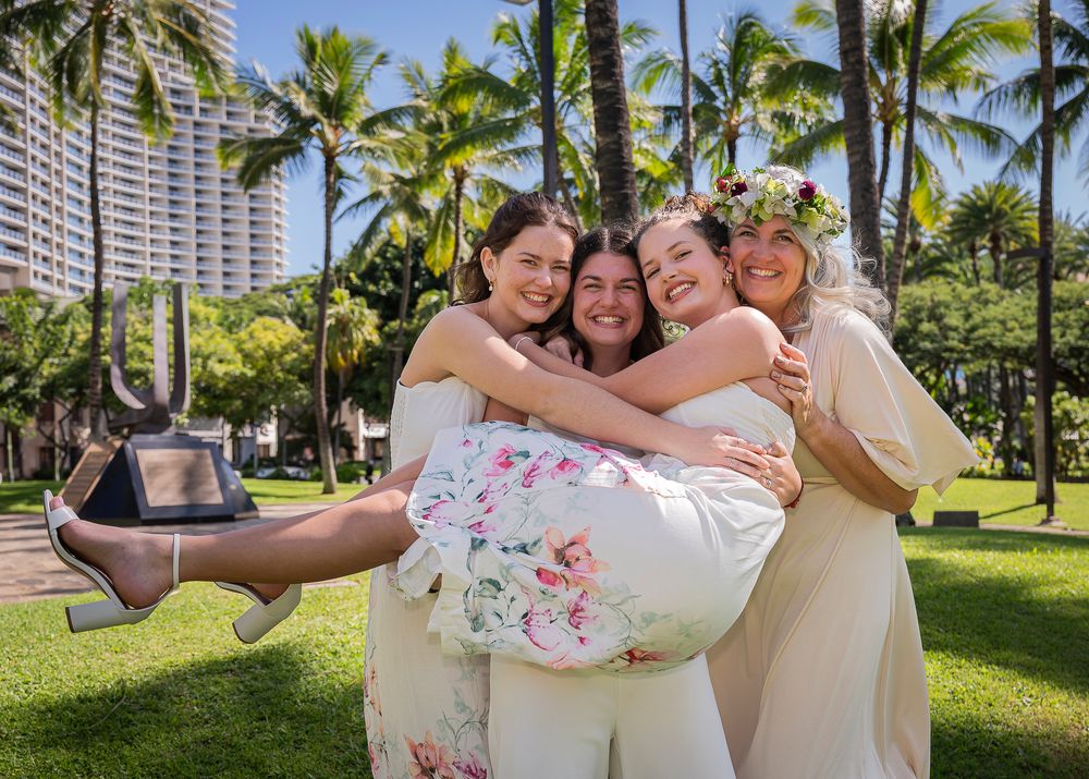 Three people in white and floral outfits embrace joyfully in a tropical garden setting with palm trees and buildings.