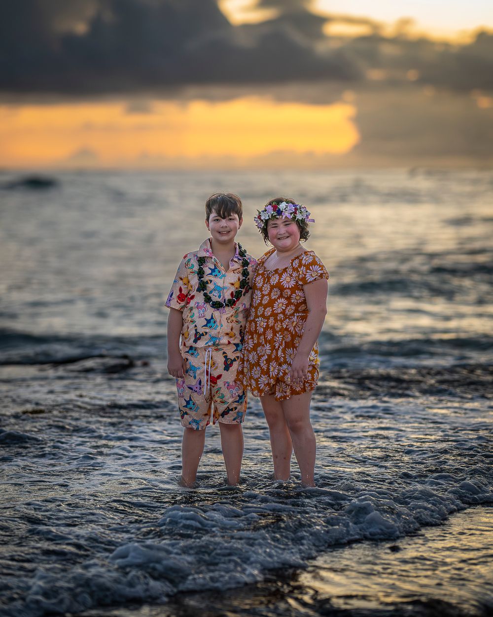 Two siblings in floral outfits stand on wet sand at sunset as waves lap at their feet on Oahu beach.