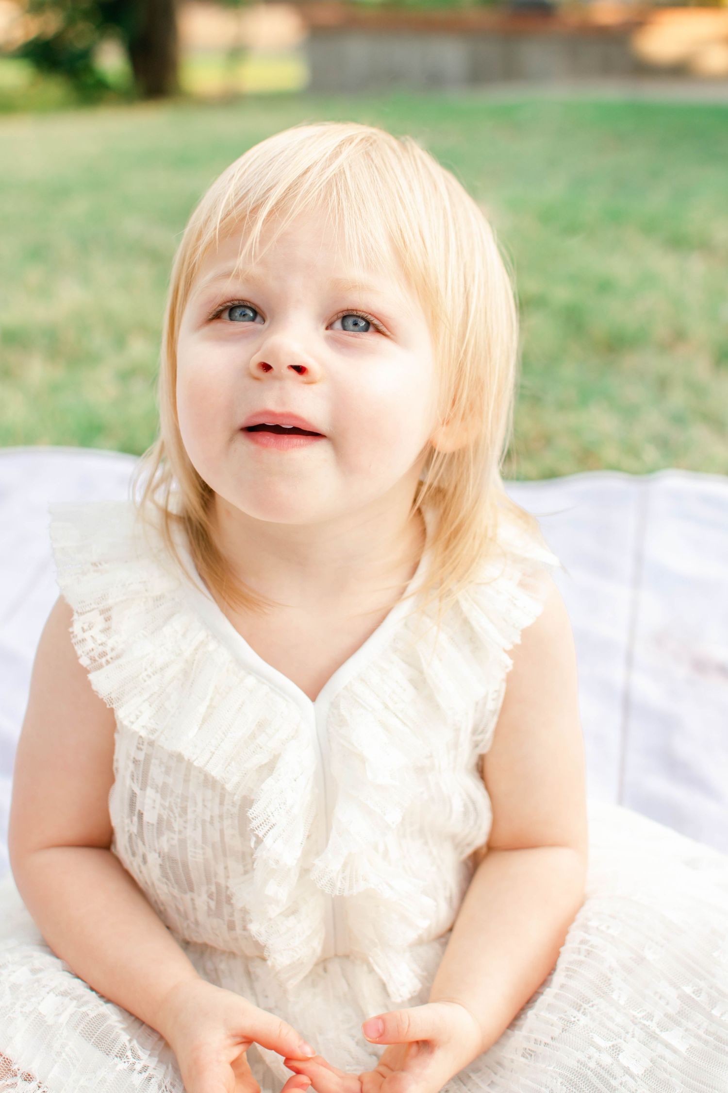 A toddler in a white lace dress lies on a blanket outdoors, looking up with joy.