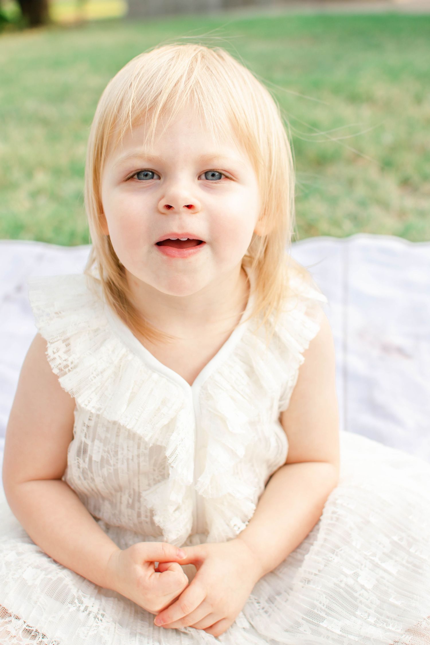 A child in a white lace dress lies on a blanket outdoors with green grass in the background.