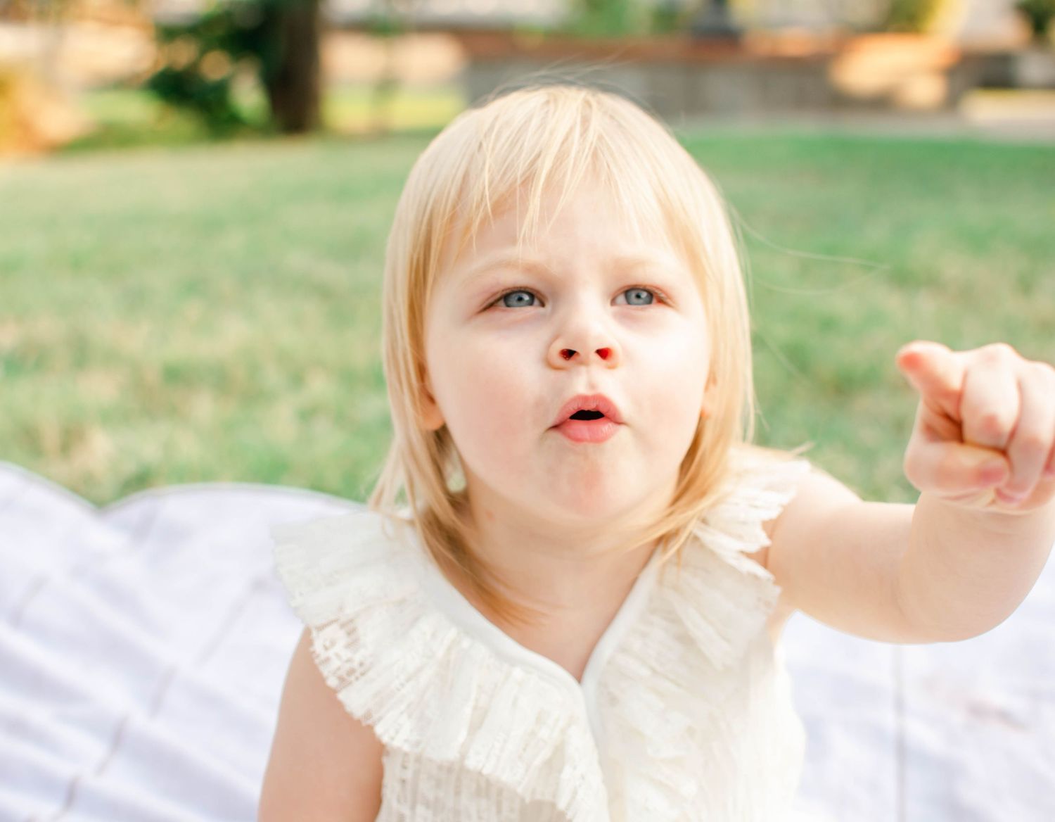 A young child in a white dress points while sitting outdoors in a sunny garden setting.