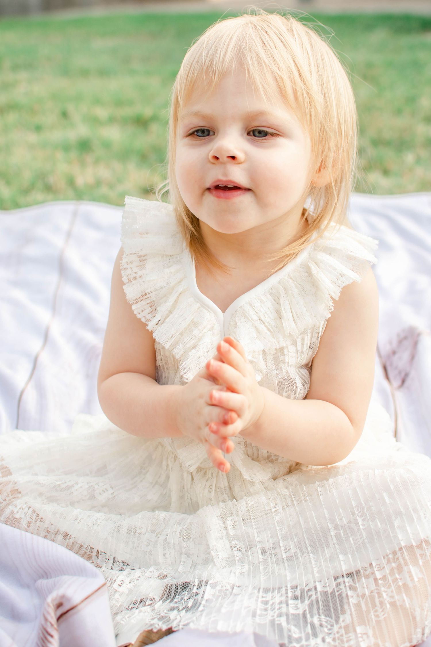 A small child in a delicate white dress plays while lying on a white blanket in the grass.