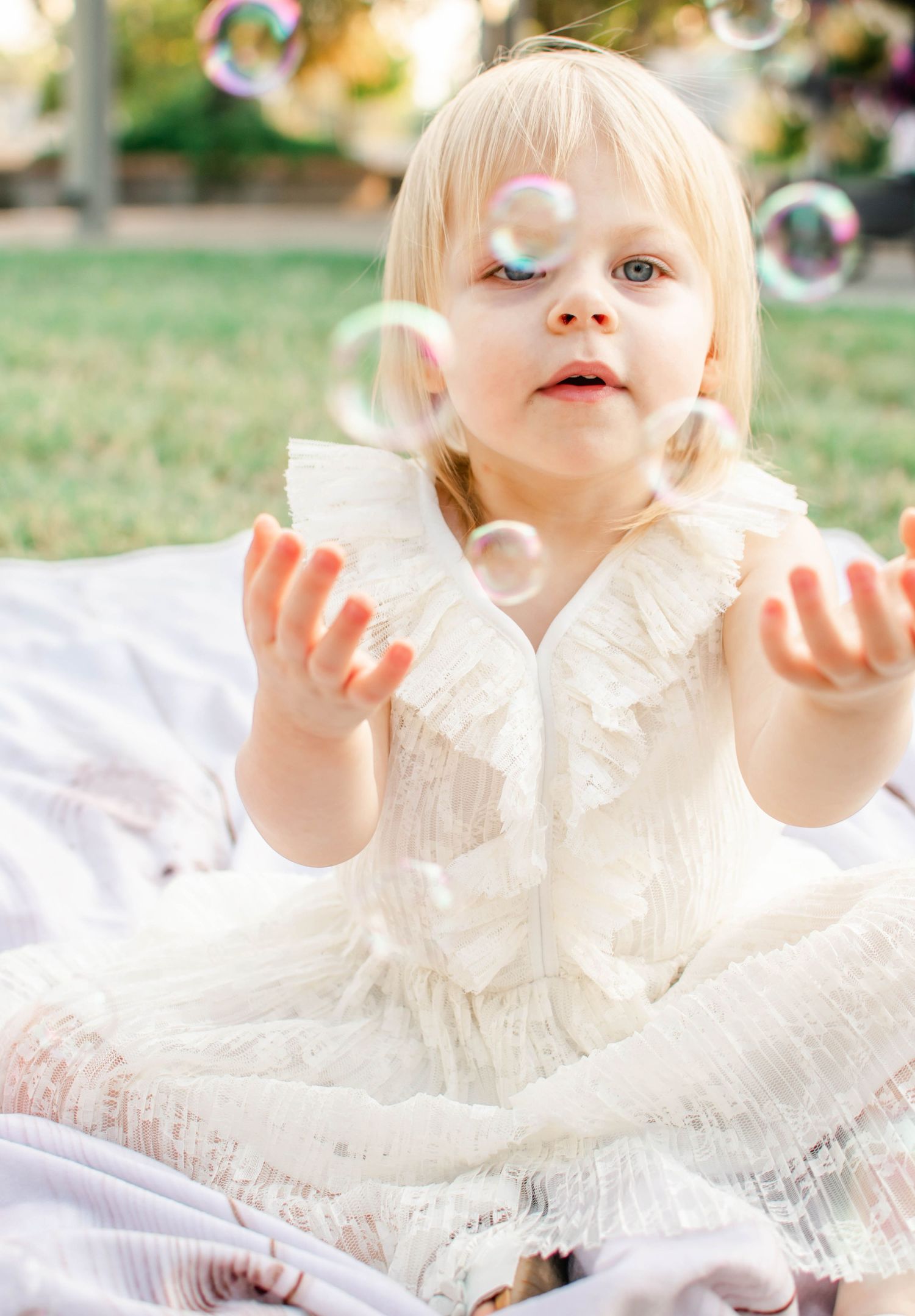 A young child in white ruffled dress reaches for bubbles while playing on a blanket outdoors.