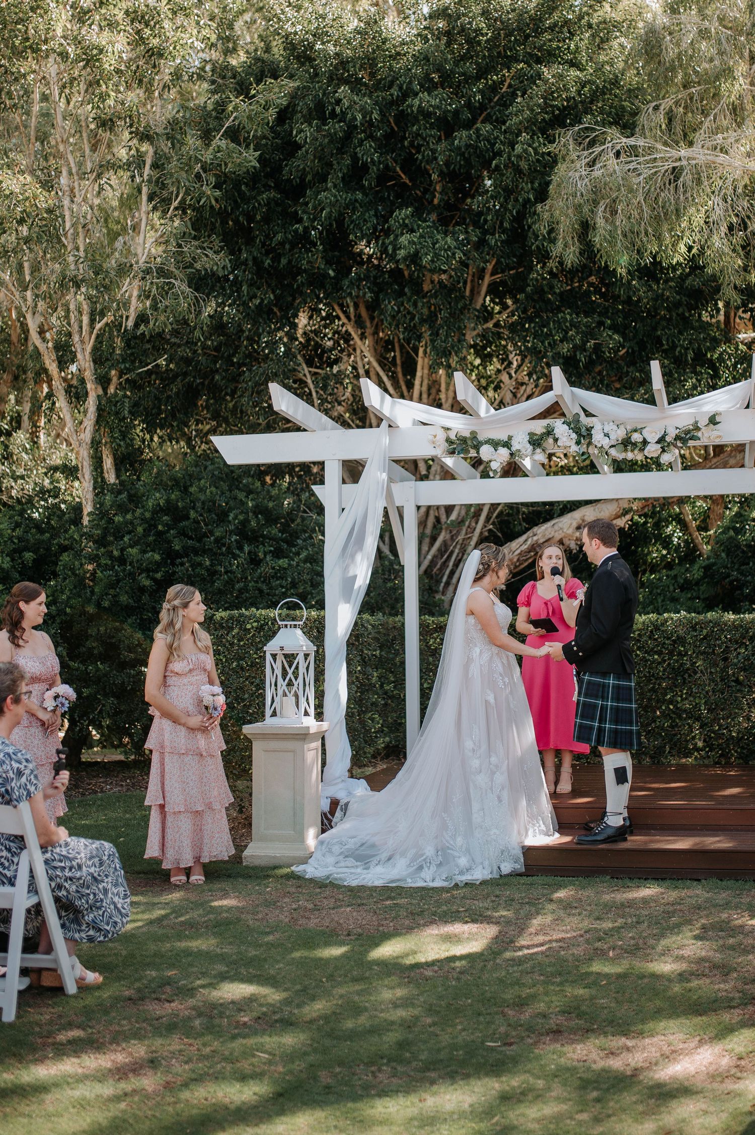 An outdoor garden wedding ceremony takes place under a white pergola decorated with flowing fabric and surrounded by trees.