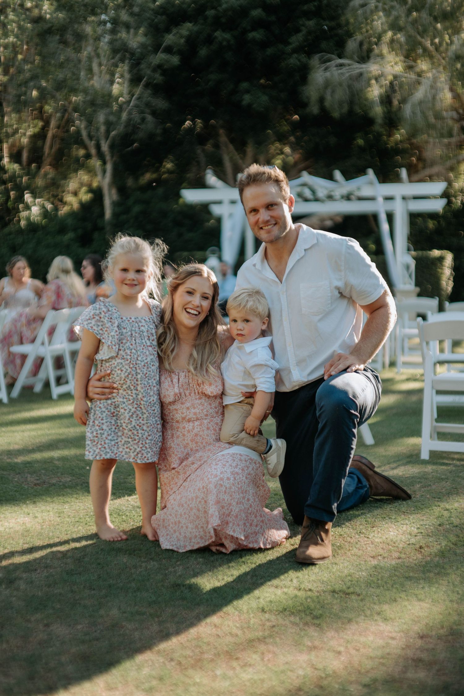 Family enjoying a playful moment together on the grass at an outdoor event with white chairs in the background.