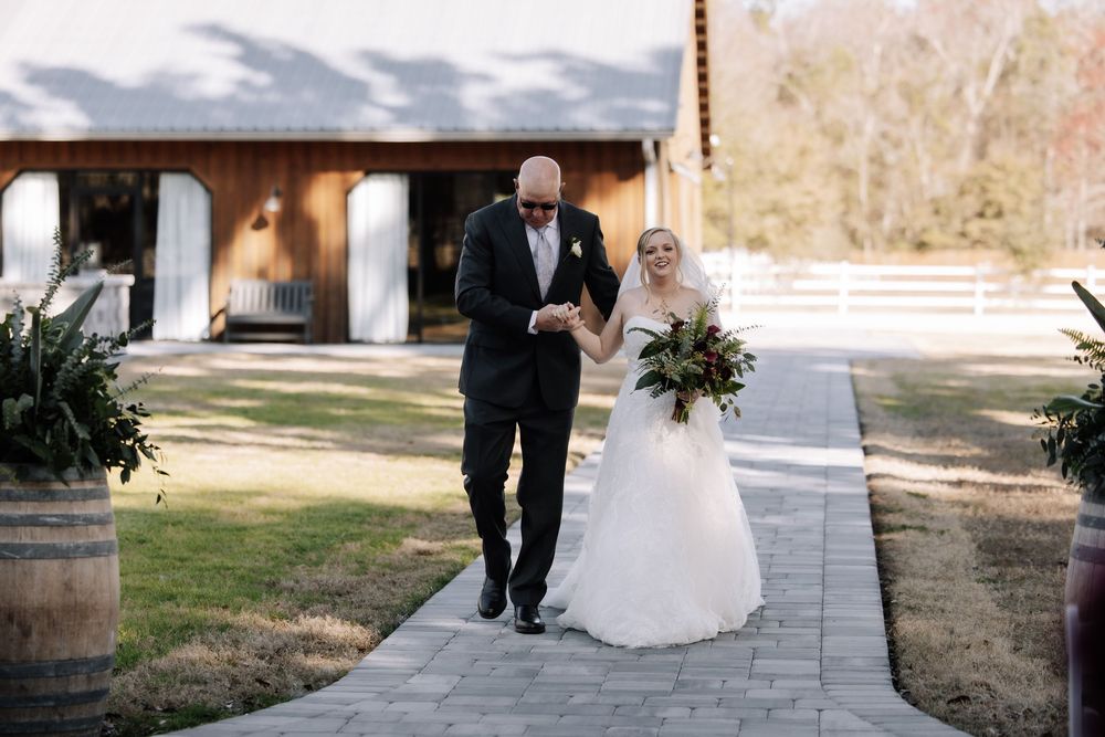 A romantic wedding couple strolls together through a rustic outdoor venue with warm sunlight filtering through trees.