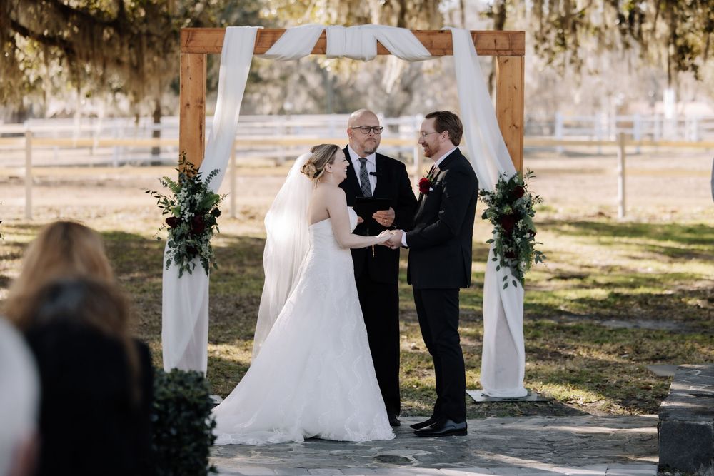A wedding ceremony takes place under a wooden arch decorated with white fabric and greenery in an outdoor setting.