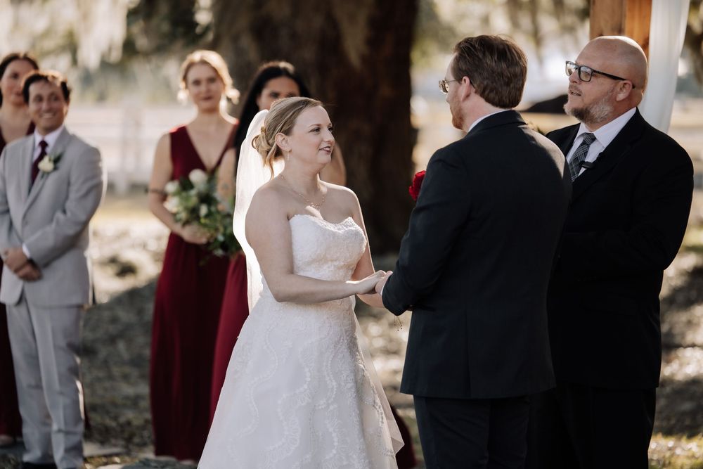 A wedding ceremony takes place outdoors with the bride in a white dress exchanging vows with the groom.