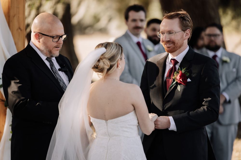 Wedding guests gather outdoors for a ceremony with people standing in formal attire under natural lighting.