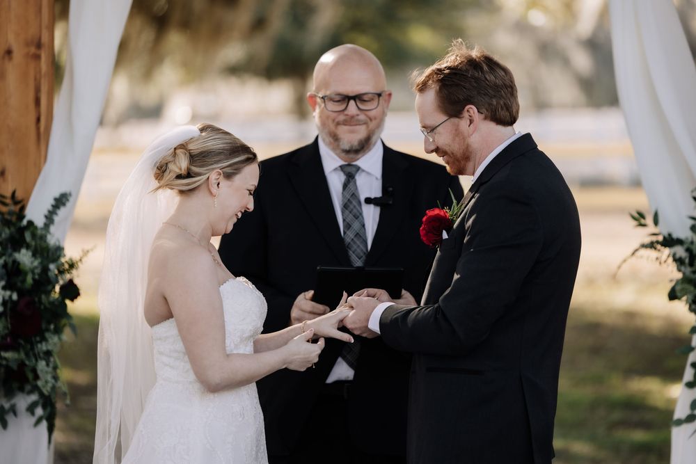 A couple exchanges rings during an outdoor wedding ceremony with an officiant presiding over the moment.
