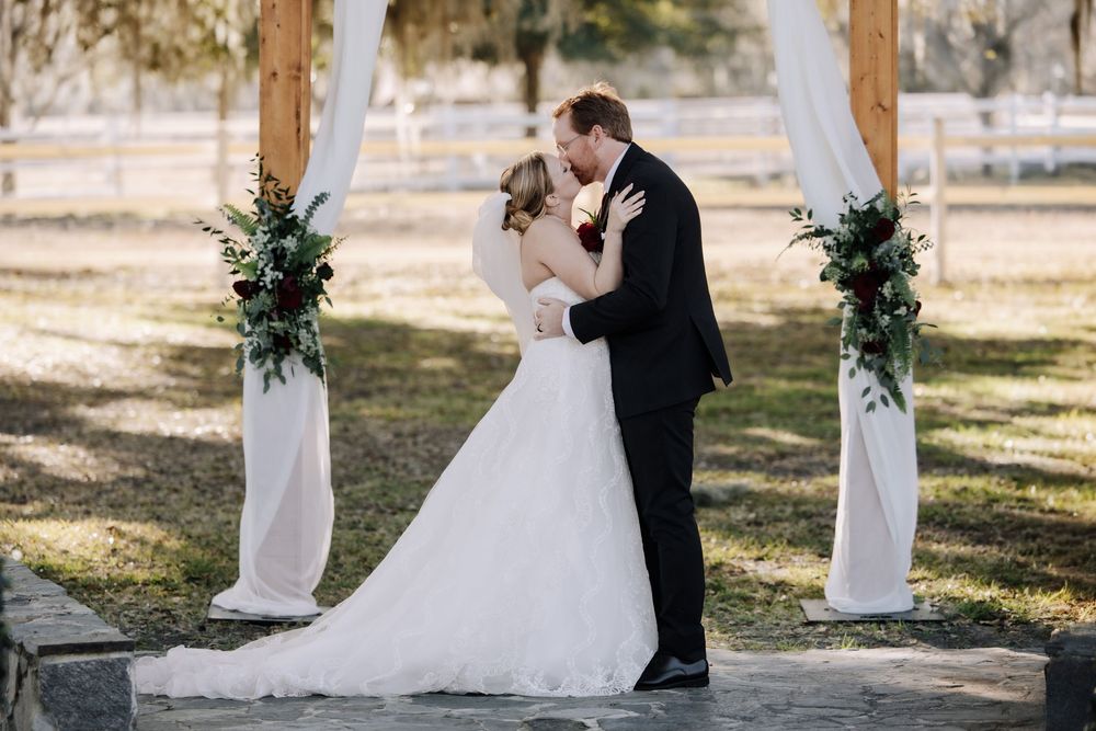 A romantic outdoor wedding ceremony under a white draped arch decorated with greenery in a sunlit garden setting.