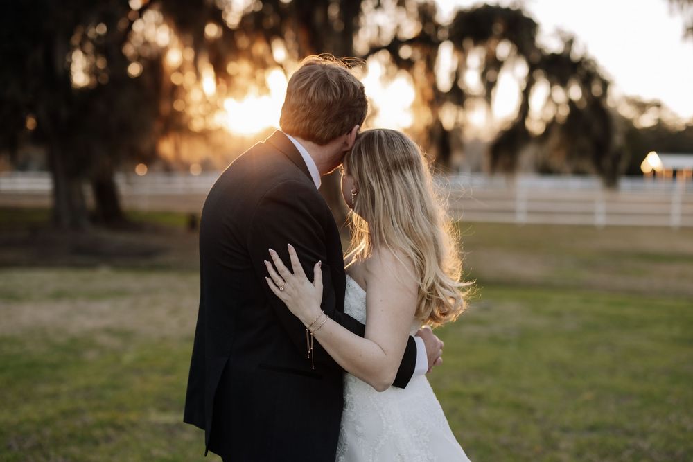 A romantic couple shares tender moments at sunset in a rustic outdoor setting with white fences and trees.