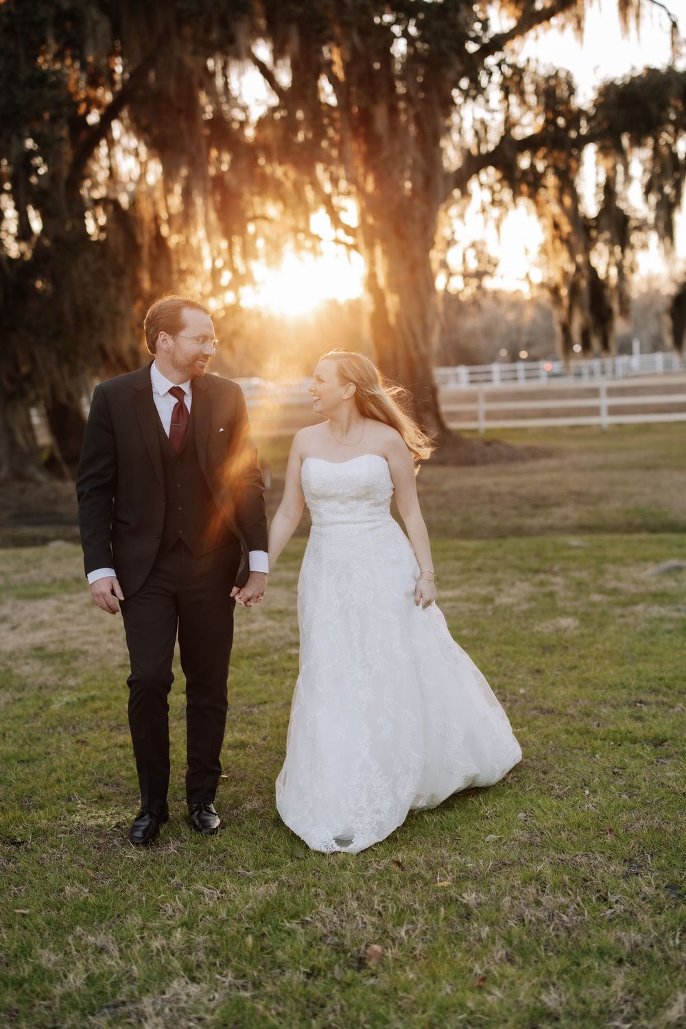 Newlyweds walk together at sunset through a field with Spanish moss draped trees creating a romantic silhouette.