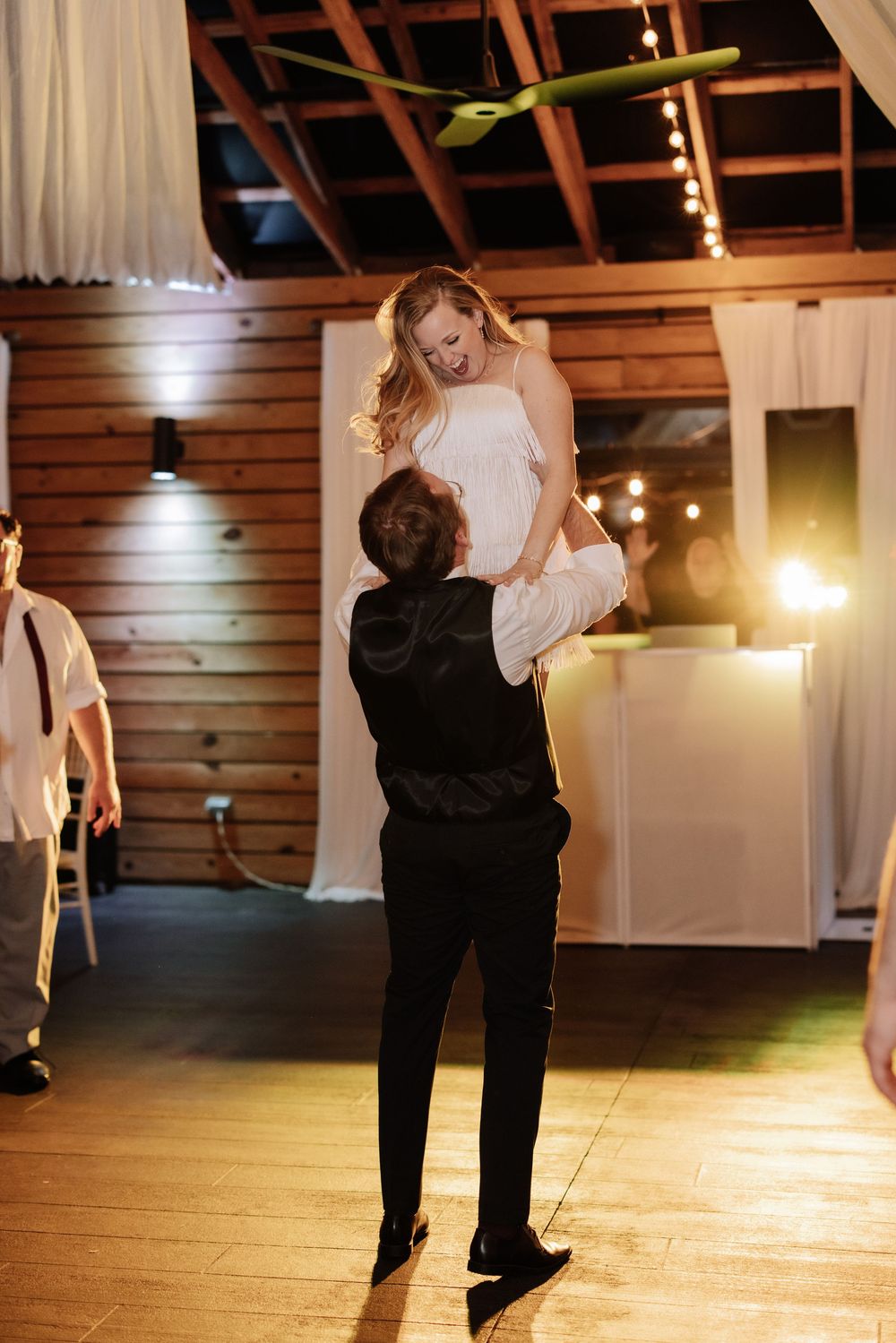 Couple dancing together in a rustic wooden barn venue illuminated by warm string lights during a special celebration.