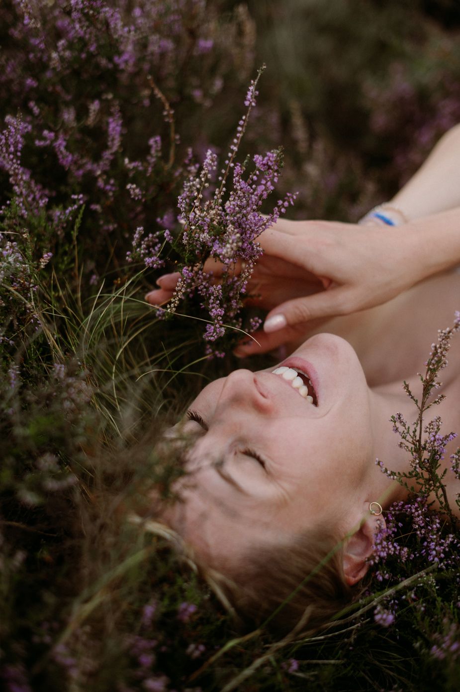 A person lies dreamily among delicate purple heather flowers in a moody outdoor setting.