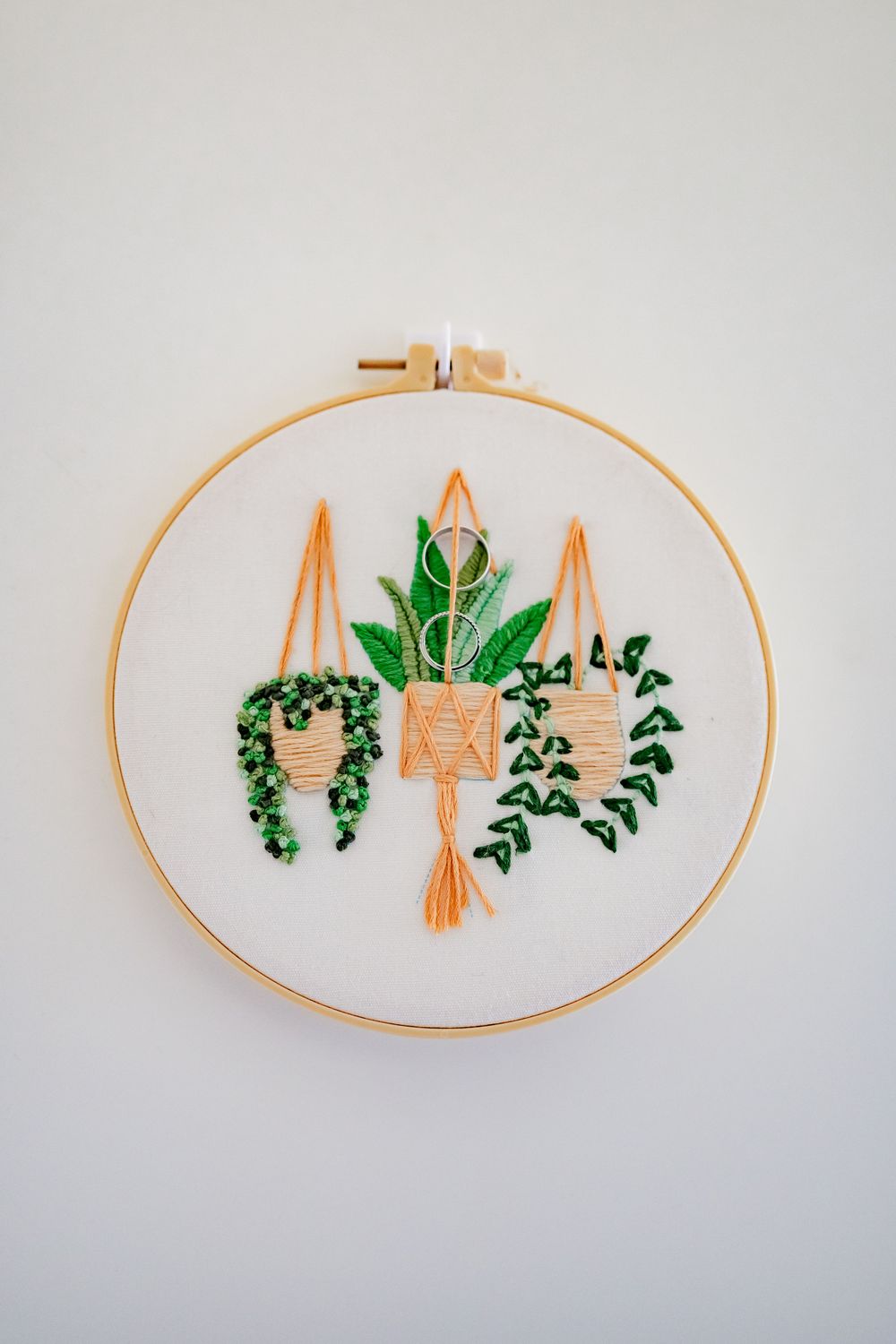 Embroidery hoop art featuring hanging plants in green and orange thread on white fabric.
