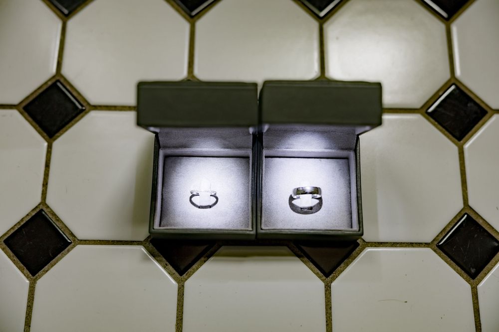 Two wedding rings in open black jewelry boxes on tiled floor.