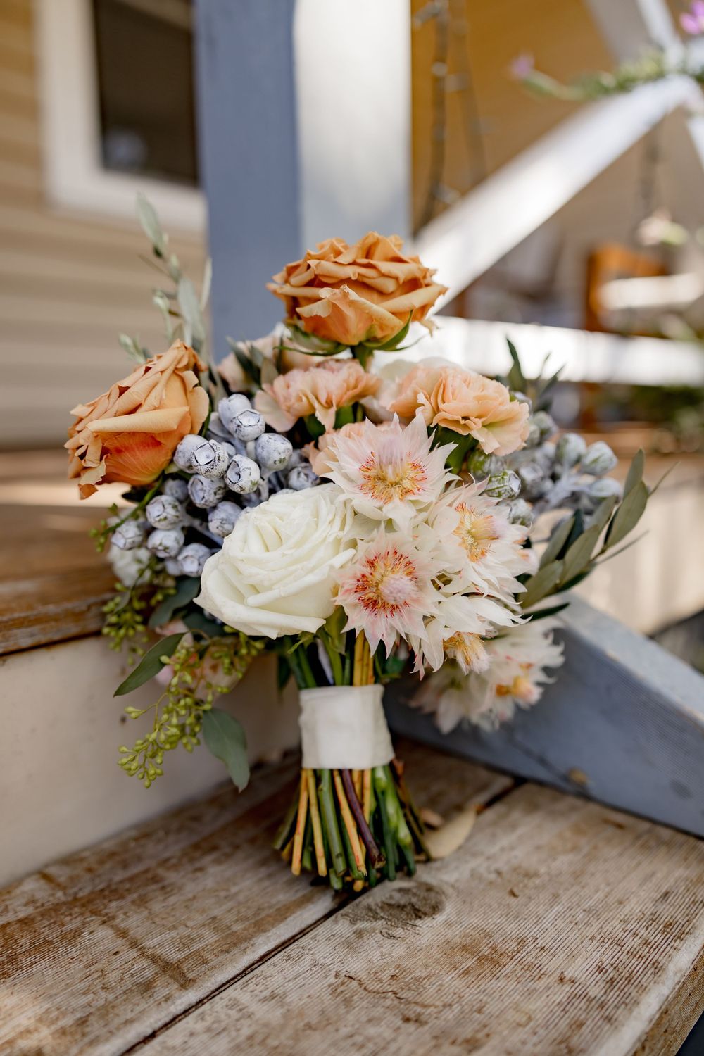 Bridal bouquet with peach roses, white flowers, and blue accents on wooden surface.