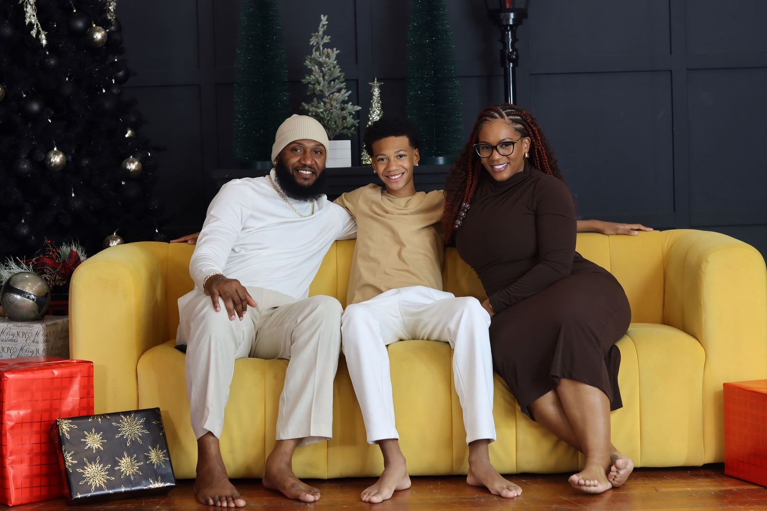 Three people sit together on a yellow couch with Christmas trees and holiday decorations in the background.
