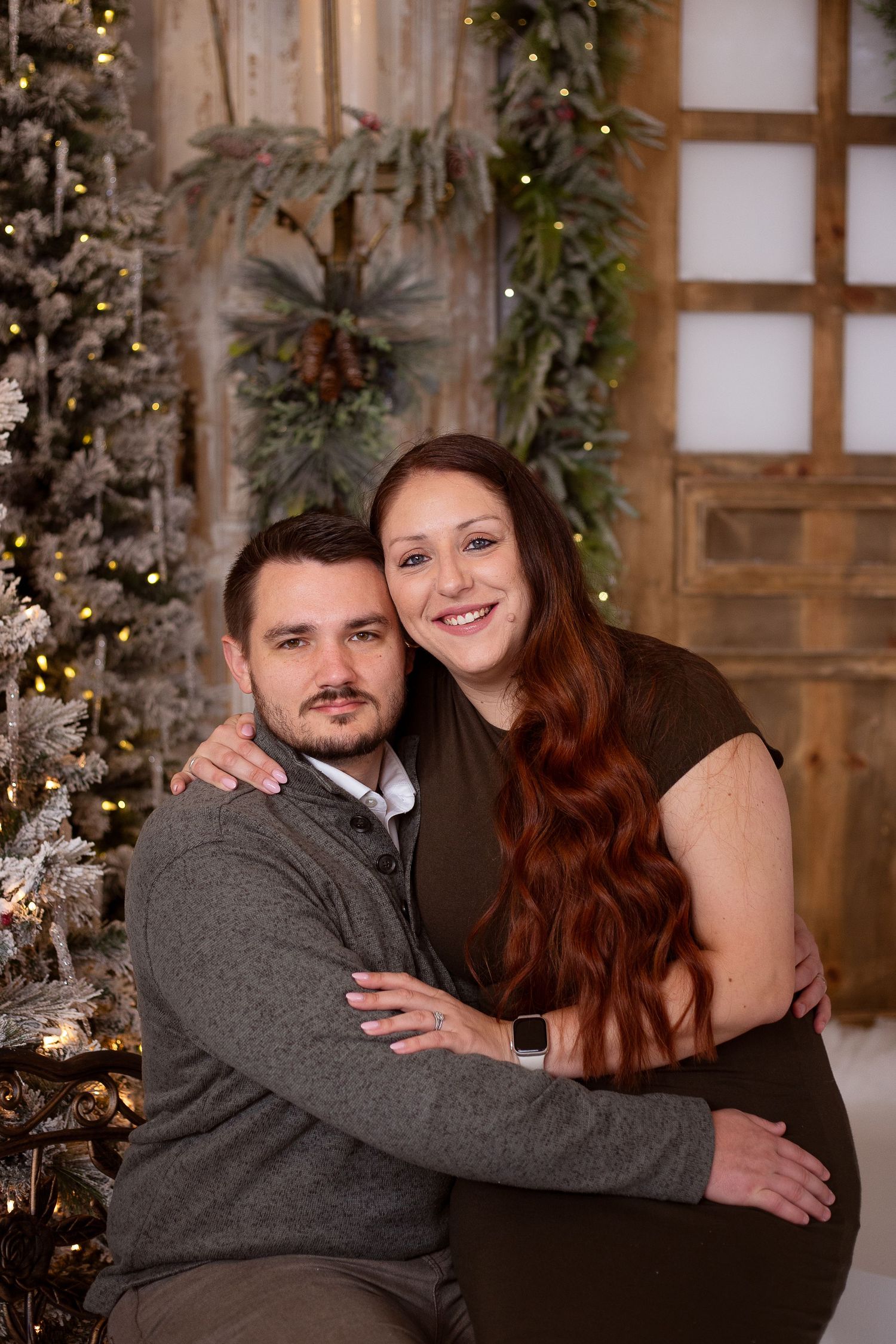 A couple poses together in front of a decorated Christmas tree and holiday greenery in a cozy indoor setting.