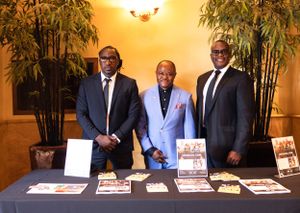 Three people in suits stand behind a display table with books and materials at an event.