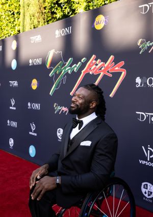 Person in wheelchair and dark suit smiles on red carpet with event backdrop.