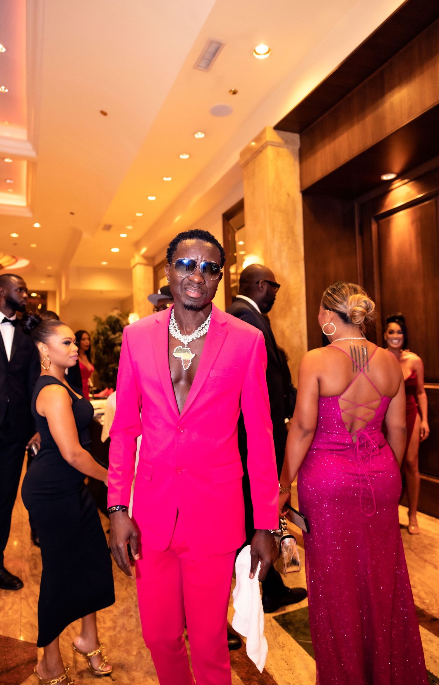 A person in a vibrant pink suit strikes a confident pose at an elegant event with guests in formal attire.