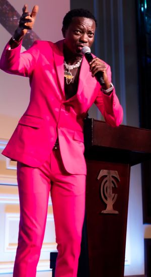 Speaker in vibrant pink suit gestures while presenting at podium with projected backdrop.