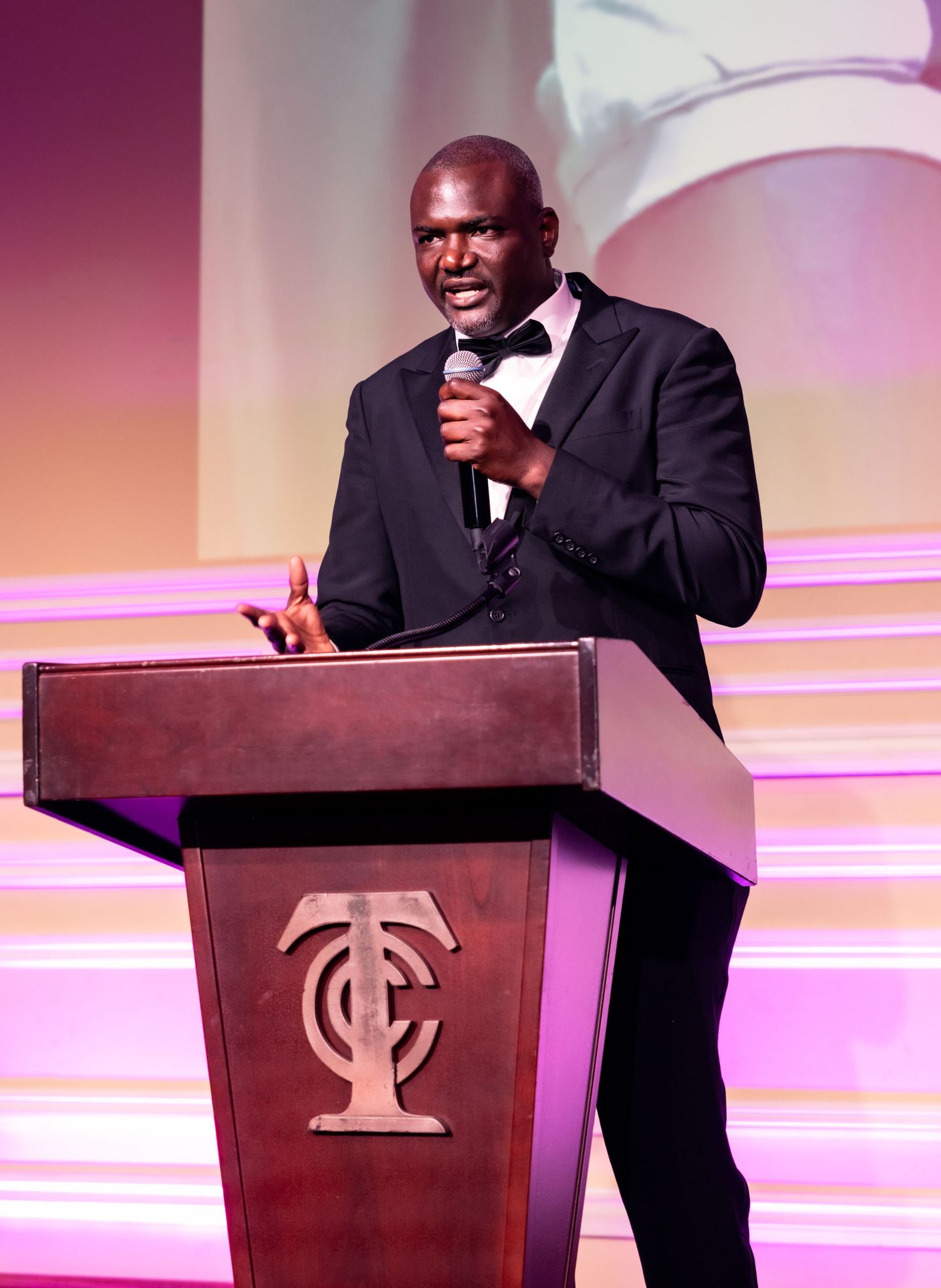 Speaker in black tuxedo delivers remarks at wooden podium with TC logo during formal event.