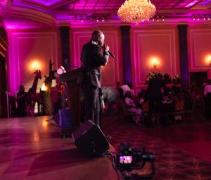 A performer on stage in formal attire under magenta lighting with a chandelier overhead.
