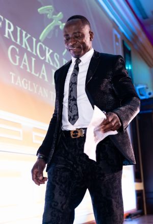 A person in a black suit and tie smiles on stage at the Afrikicks Gala event.
