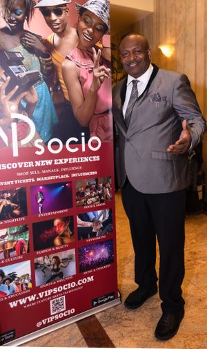 A person in a gray suit poses next to a VIP Socio event banner displaying entertainment experiences.