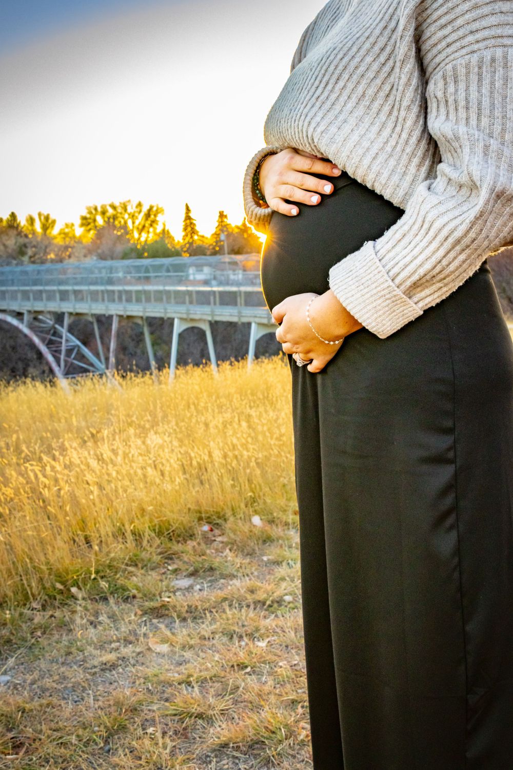 a pregnant woman holding her baby bump near a bridge.